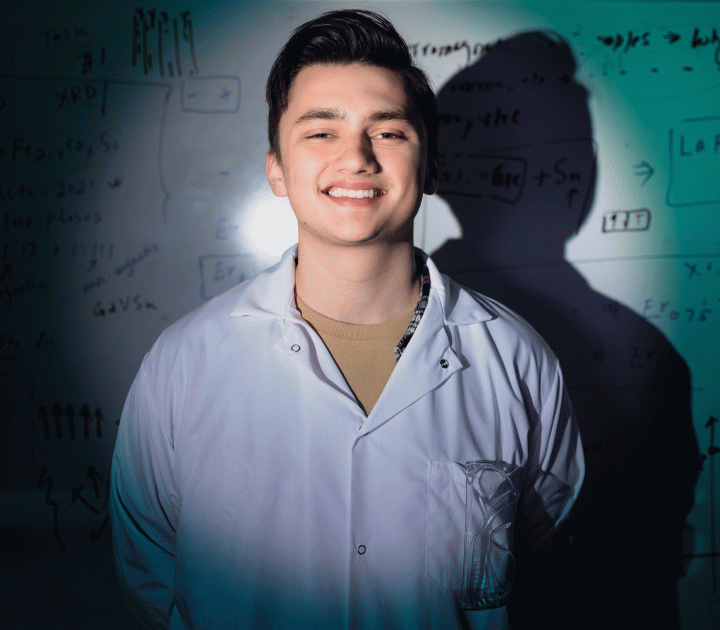 Physics sophomore Chris Burgio wearing a white lab coat and smiling in front of a white board filled with equations