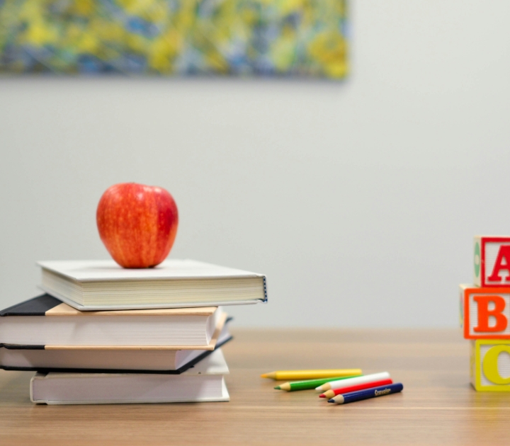 A stack of books with an apple on top, some colored pencils, and blocks reading ABC on a table