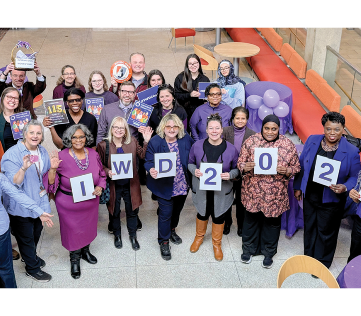 A group of male and female faculty and staff members, including interim president Bonita Durand, wearing purple and holding a sign that spells IWD 2026