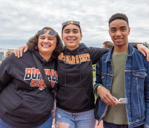 Three people donning Buffalo State sweatshirts smile for the camera on eclipse day