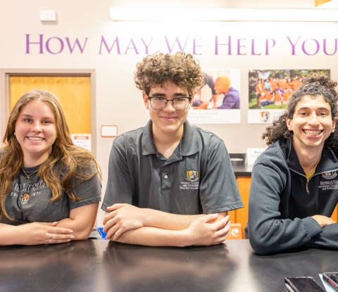 Three student orientation leaders, smiling and seated at the Information Desk. The words "How May We Help You?" are posted on the wall behind them.