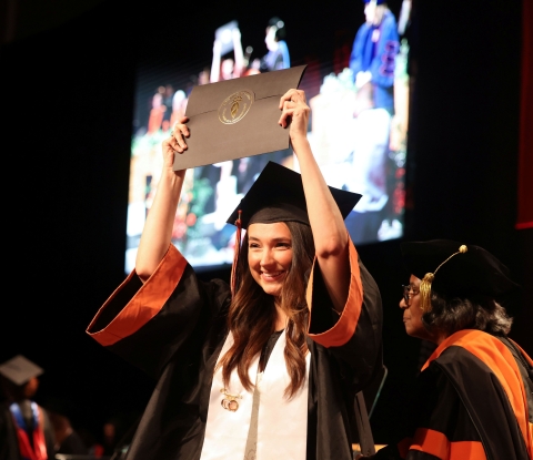 Female student in graduation cap and gown raising her diploma above her head