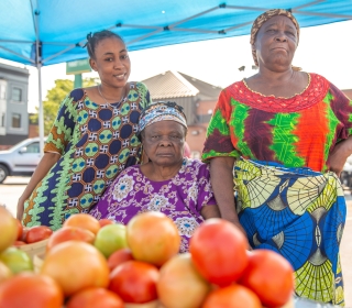 Three women vendors dressed in colorful clothing posing behind a pile of tomatoes