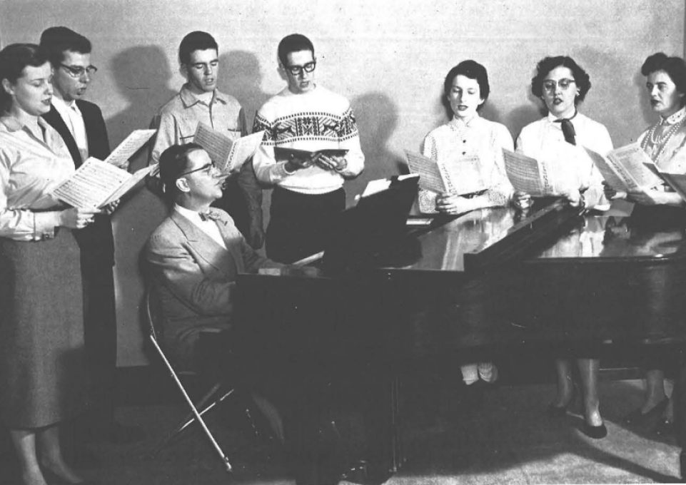 Black and white photo from1956 Elms yearbook of man playing piano and students standing and singing