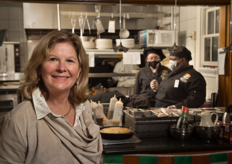 Kathleen O'Brien standing in front of the Campus House kitchen, where chefs are busy working