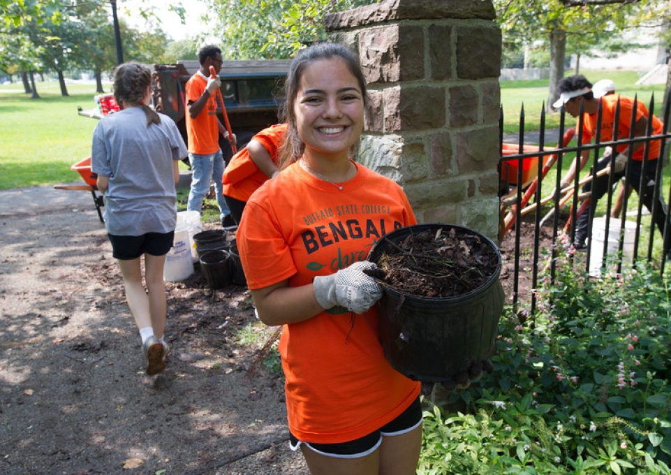 A smiling Buffalo State volunteer wearing an orange Bengals shirt and carrying a bucket of yard debris