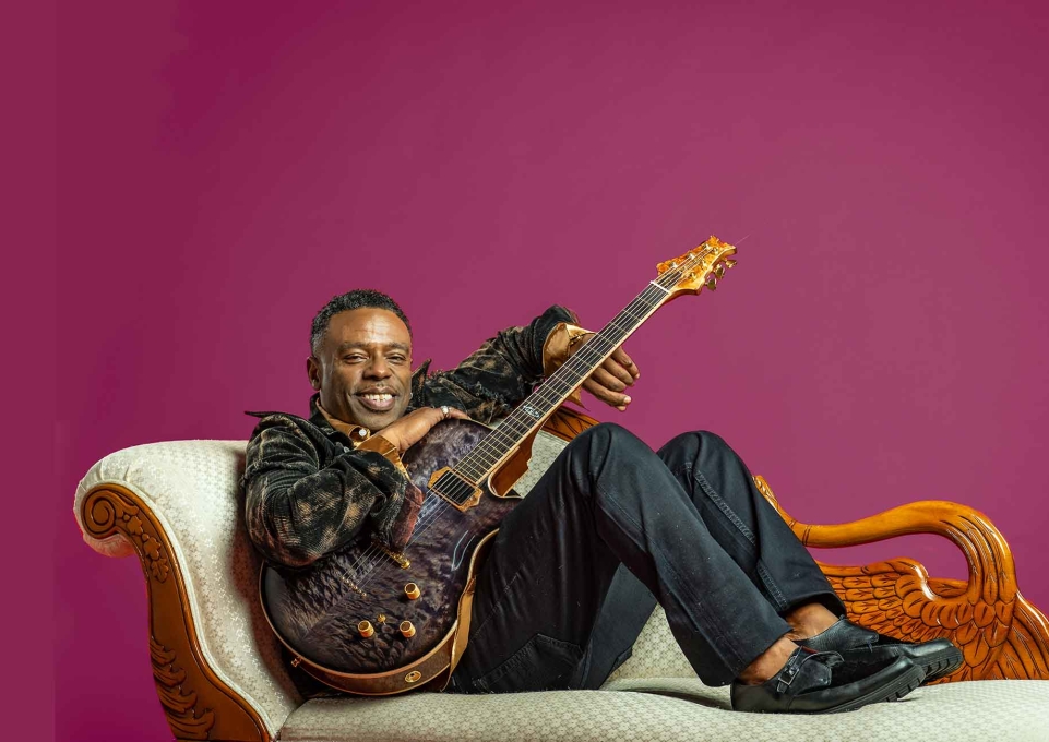 Norman Brown holding a guitar and reclining on a chaise longue