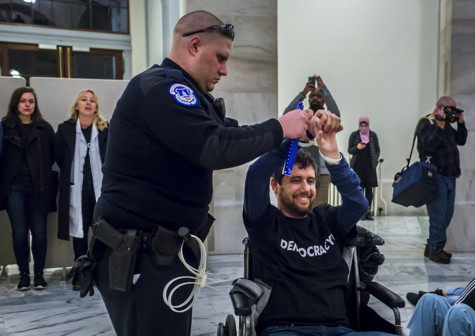 Ady Barkan in his wheelchair smiling while being handcuffed by police officer