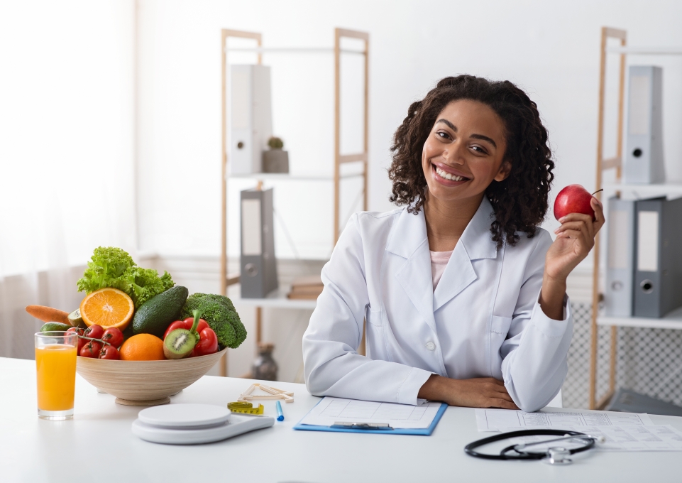 Smiling woman in a lab coat sits before a table with a bowl of fruits and vegetables, a stethoscope, and an apple