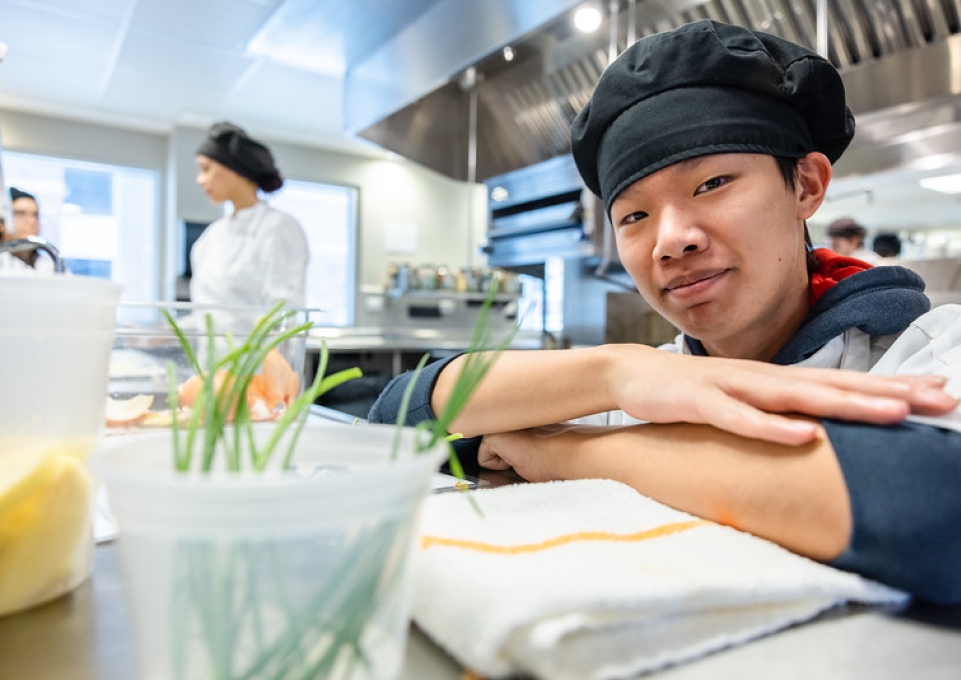 Hospitality student in chef's cap with arms crossed smiles at the camera