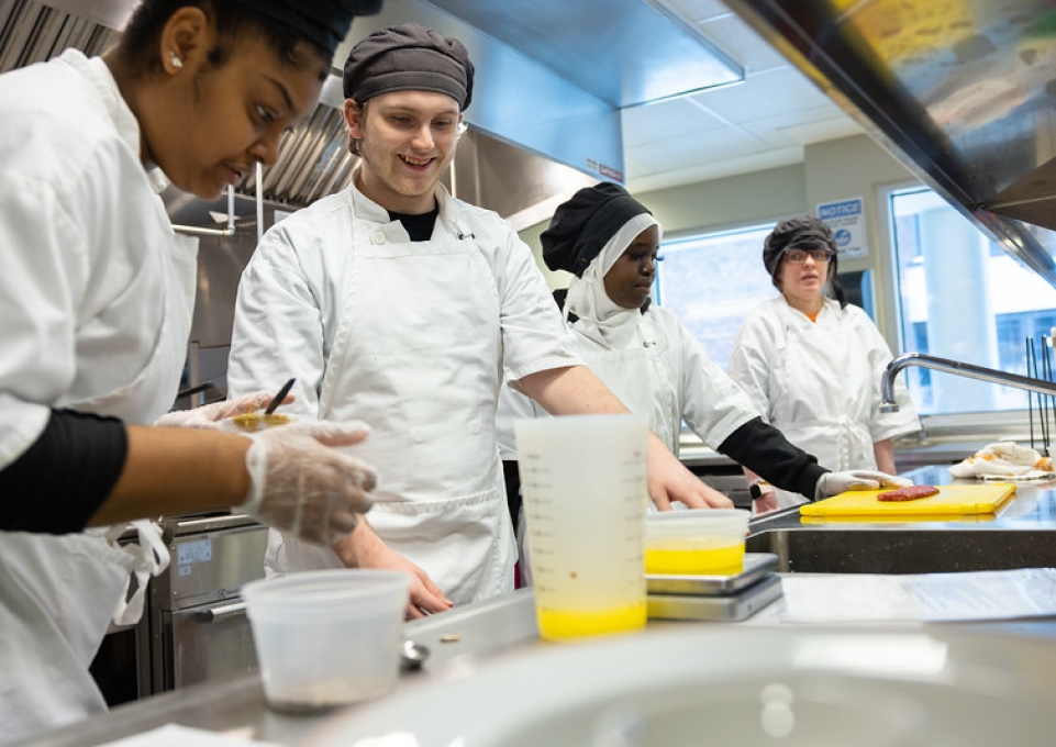 Four students wearing white chef's aprons cooking side by side